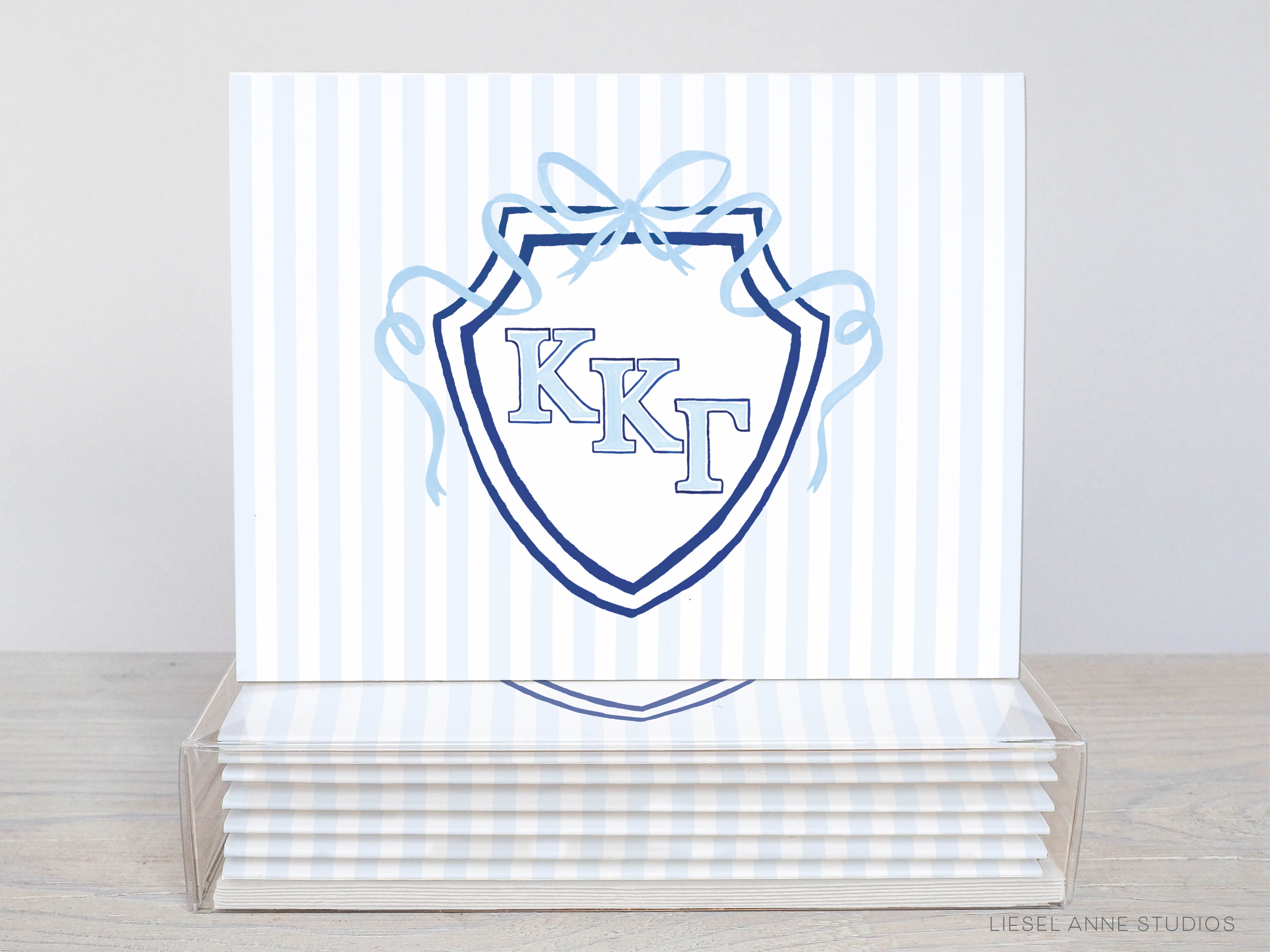 A stack of white greeting cards with a blue and white striped design and a shield-shaped logo featuring the letters "KKT" in the center.