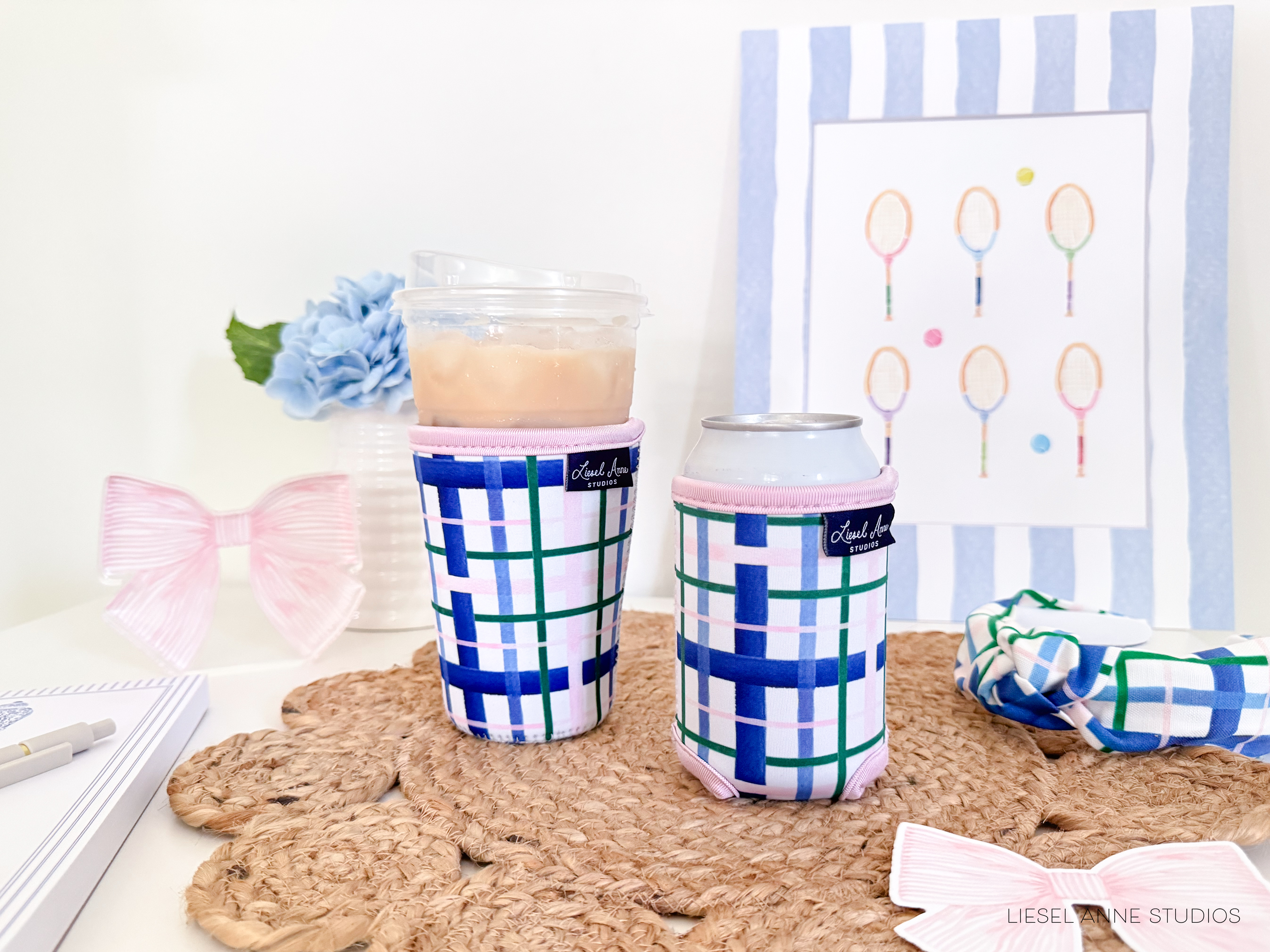 two colorful drink holders with a checkered pattern, placed on a woven mat. In the background, there is a framed picture with tennis rackets on it, and a vase with a blue flower.