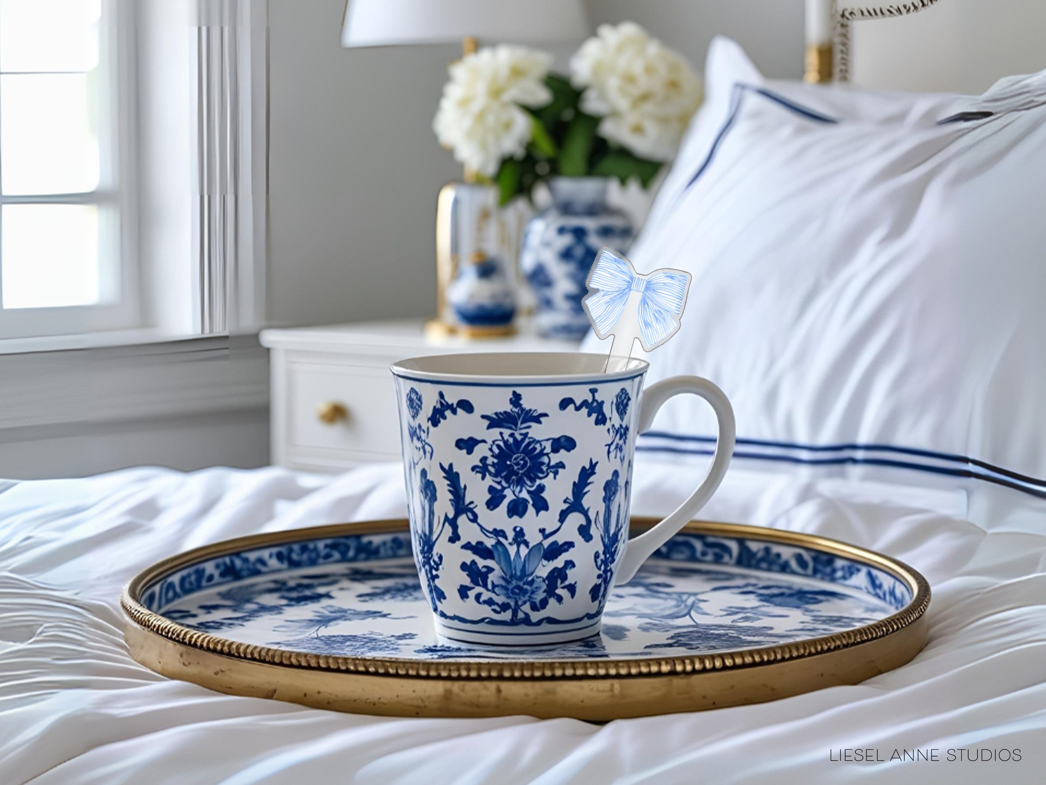 A blue and white patterned coffee mug with a butterfly-shaped stirrer sits on a white tray, placed on a bed with white sheets and a white pillow.