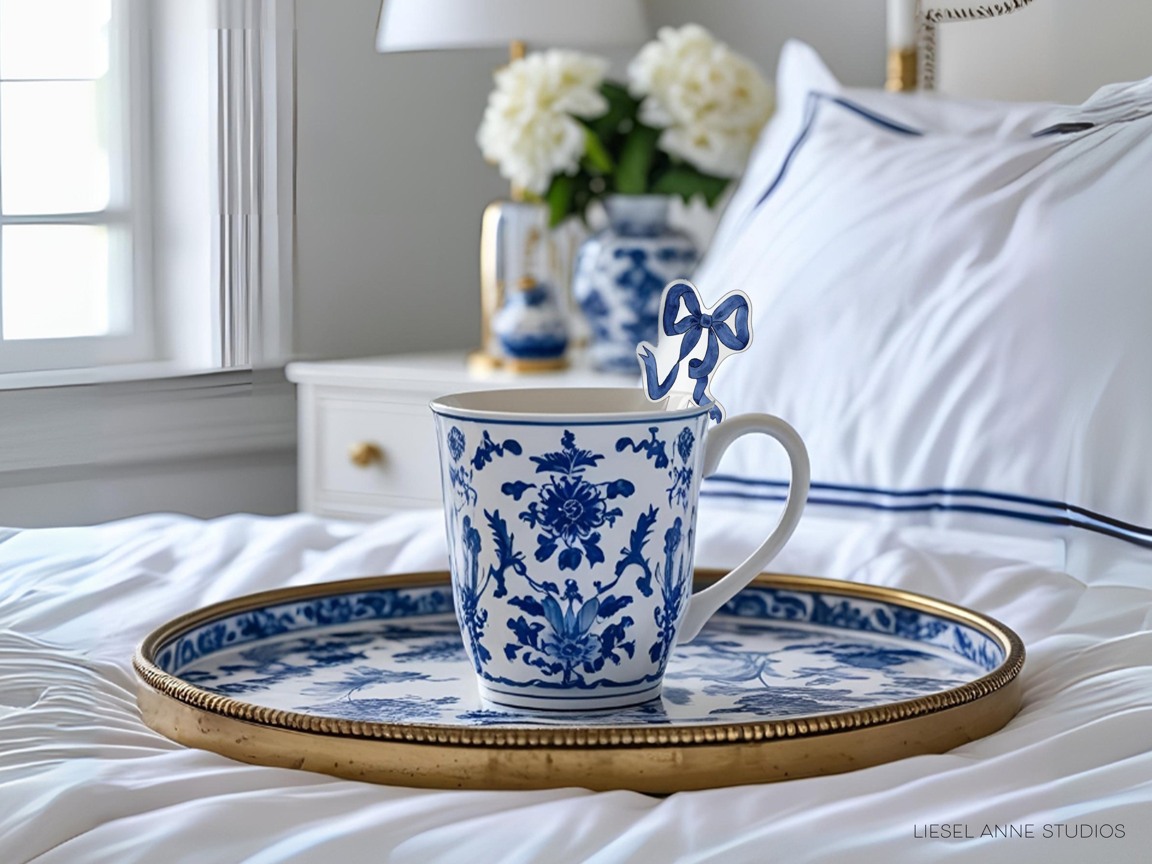A blue and white patterned coffee mug with a bow on top rests on a gold tray, placed on a bed with white sheets and a white pillow.