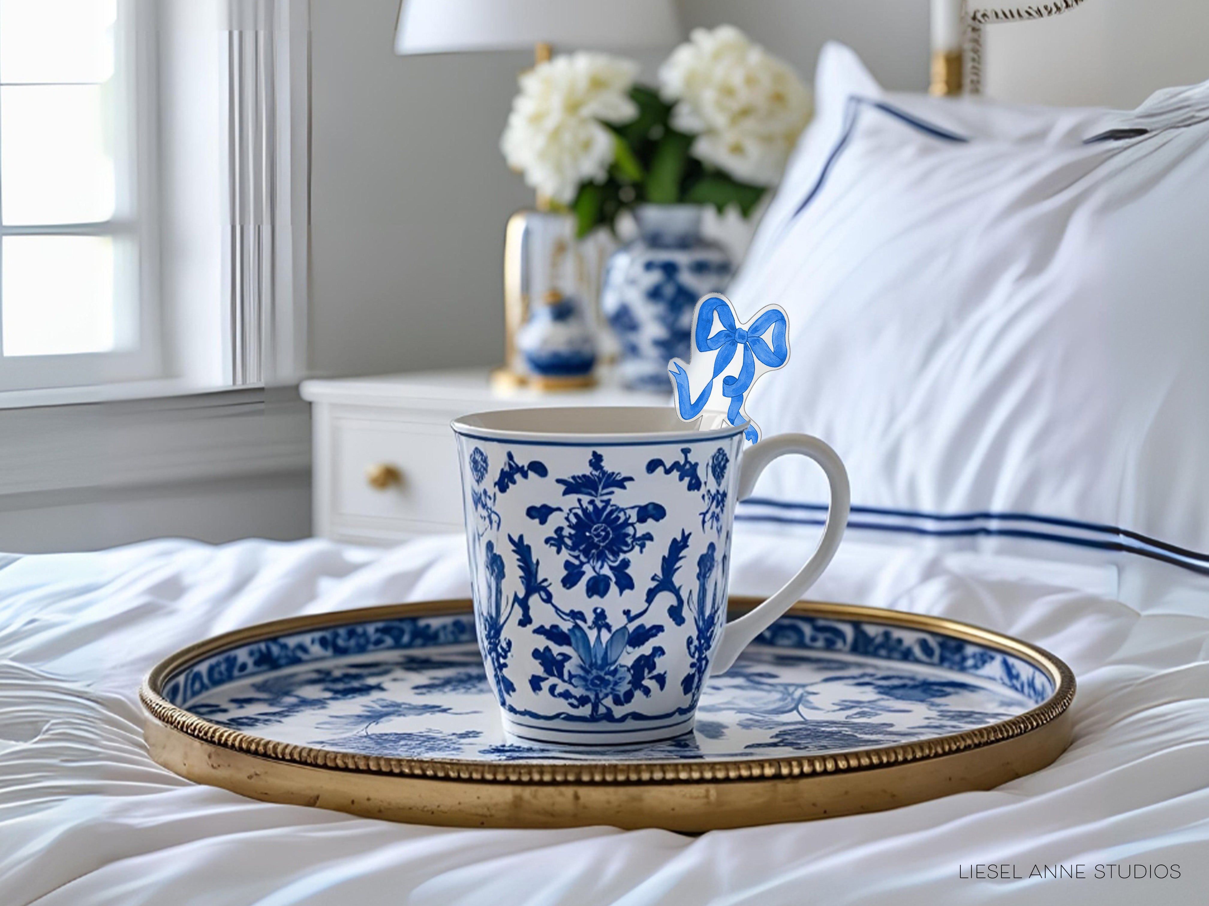 A blue and white patterned coffee mug with a bow on top rests on a gold tray, placed on a bed with white sheets and a white pillow.