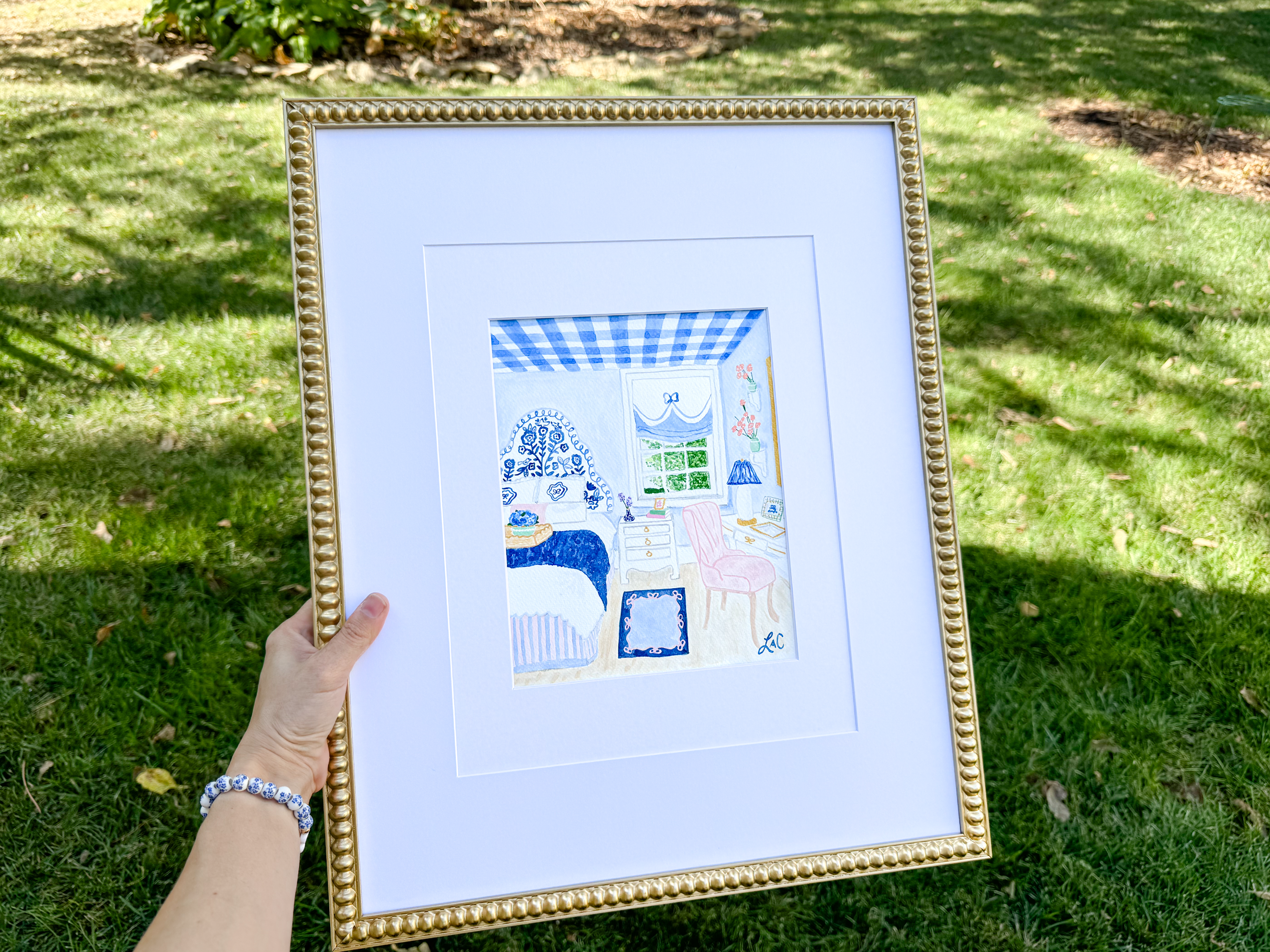 A hand holding a framed painting of a bedroom scene with a blue and white checkered bedspread, a pink chair, and a blue and white striped rug.