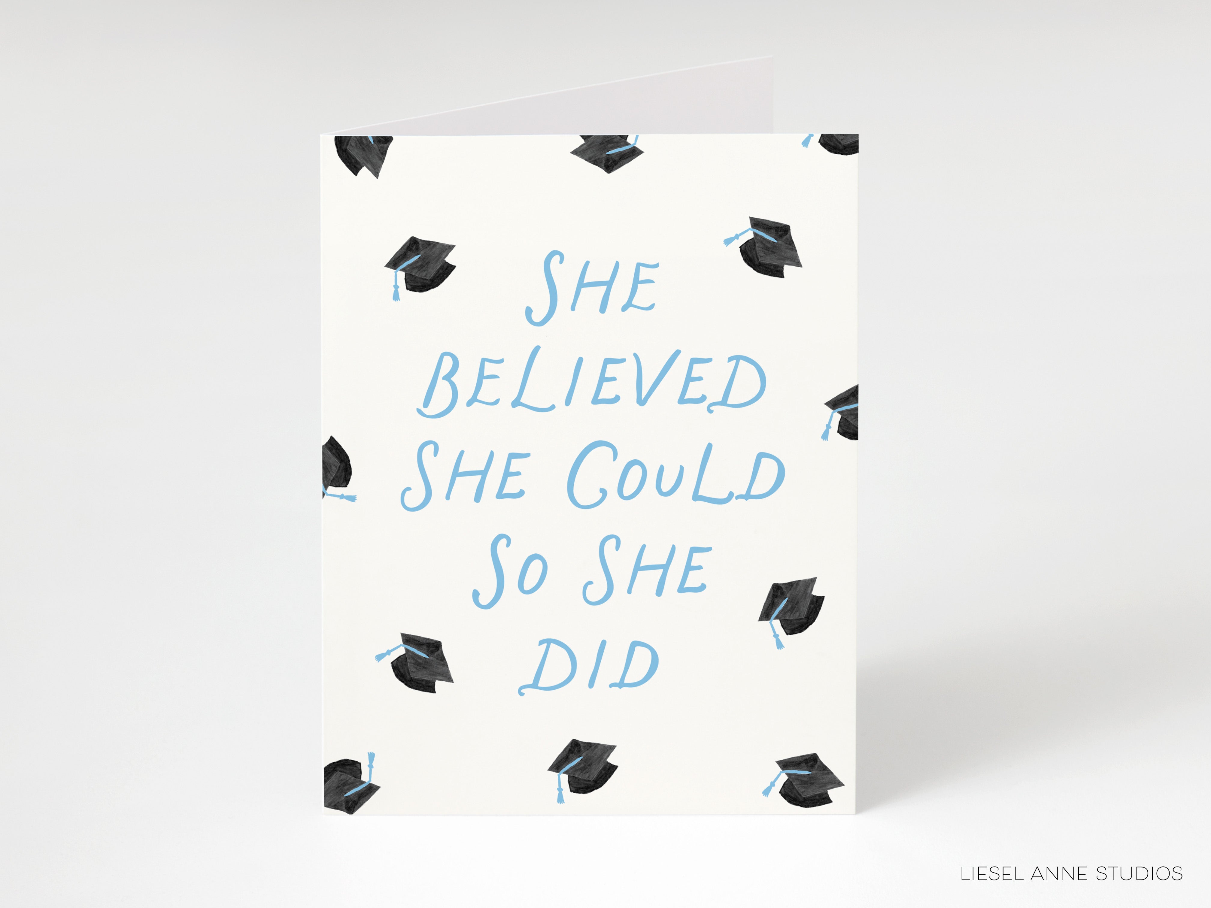 A greeting card with a white background and a pattern of black graduation caps. The text on the card reads "She believed she could so she did".