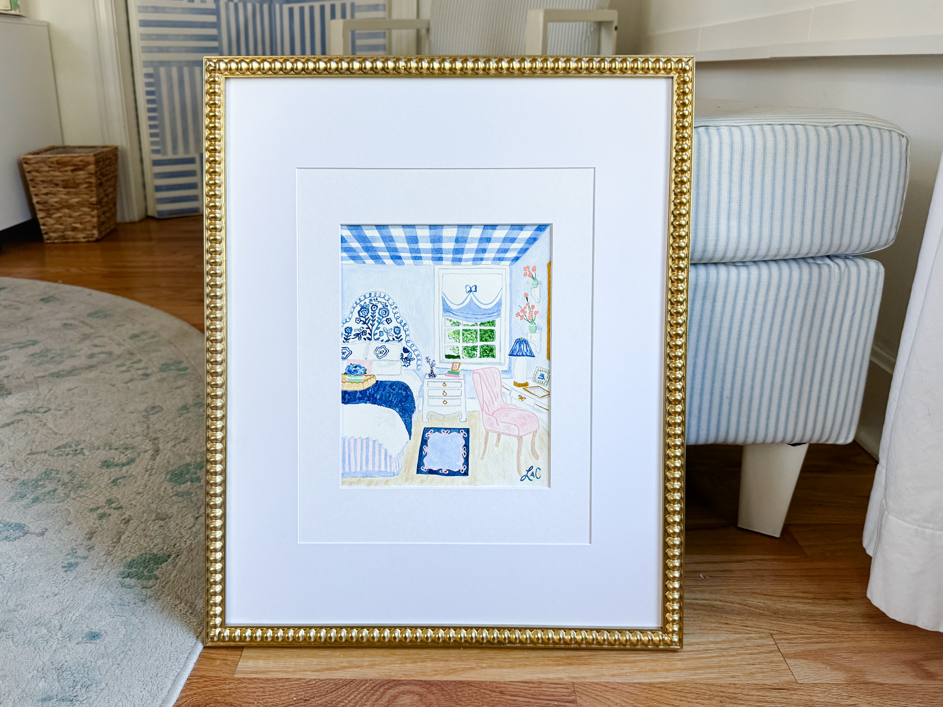 a framed painting of a bedroom scene with a blue and white checkered bedspread, a pink chair, and a blue and white striped ottoman.