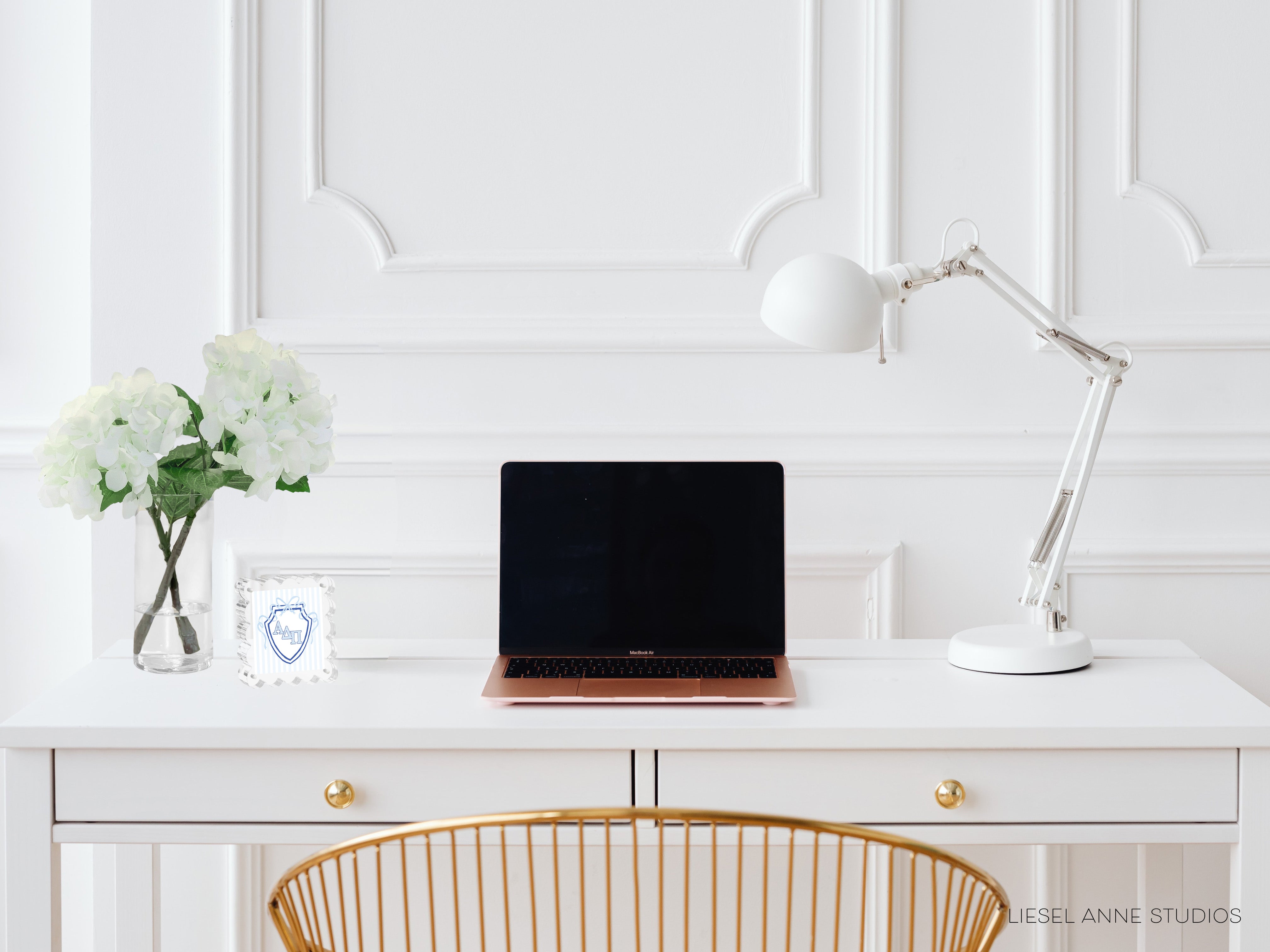 A white desk with a laptop, a vase of white flowers, and a white lamp, all arranged neatly and creating a clean, minimalist workspace.