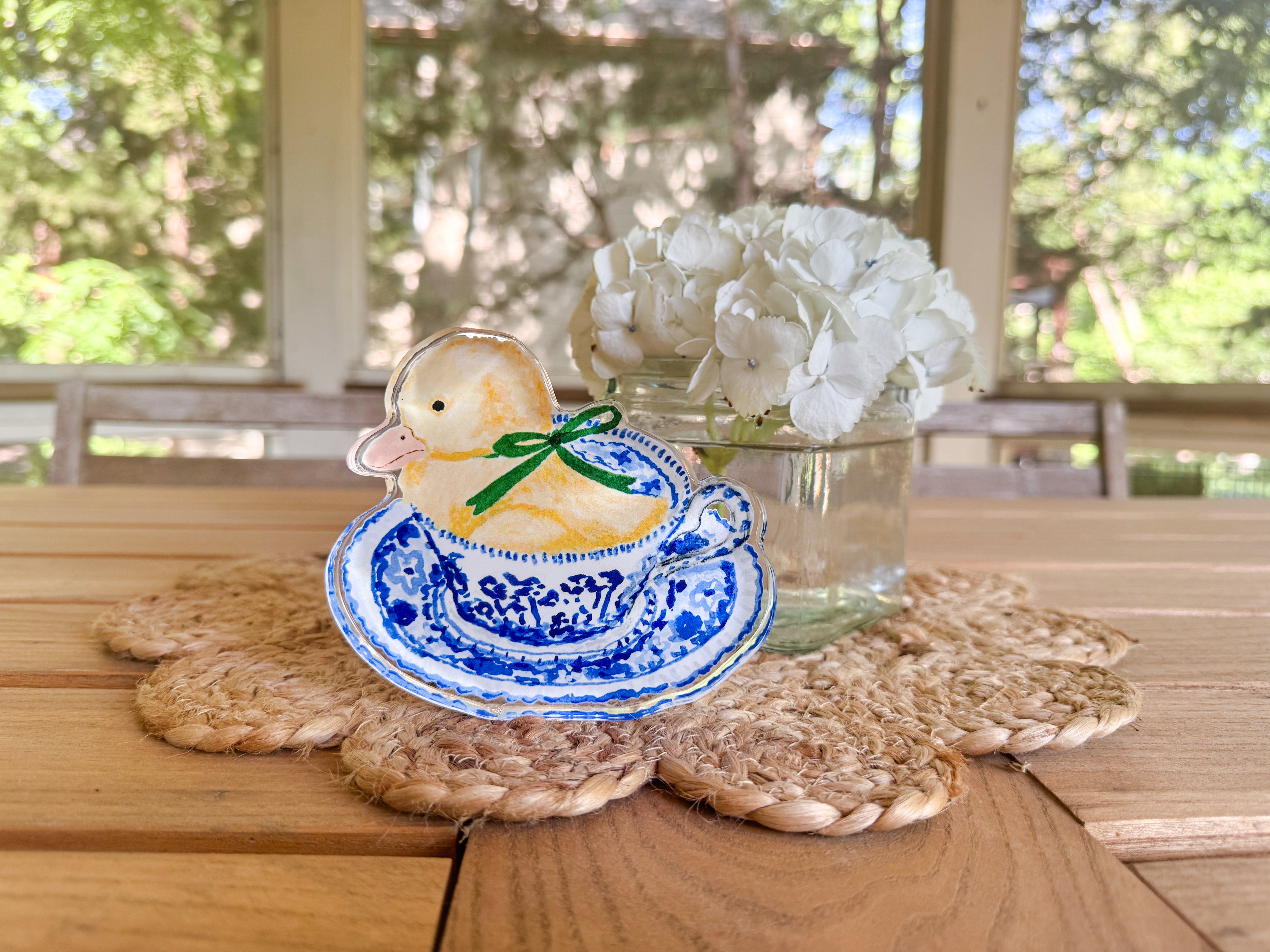 A ceramic duck figurine sitting in a blue and white teacup, placed on a woven placemat, with a vase of white hydrangeas in the background.