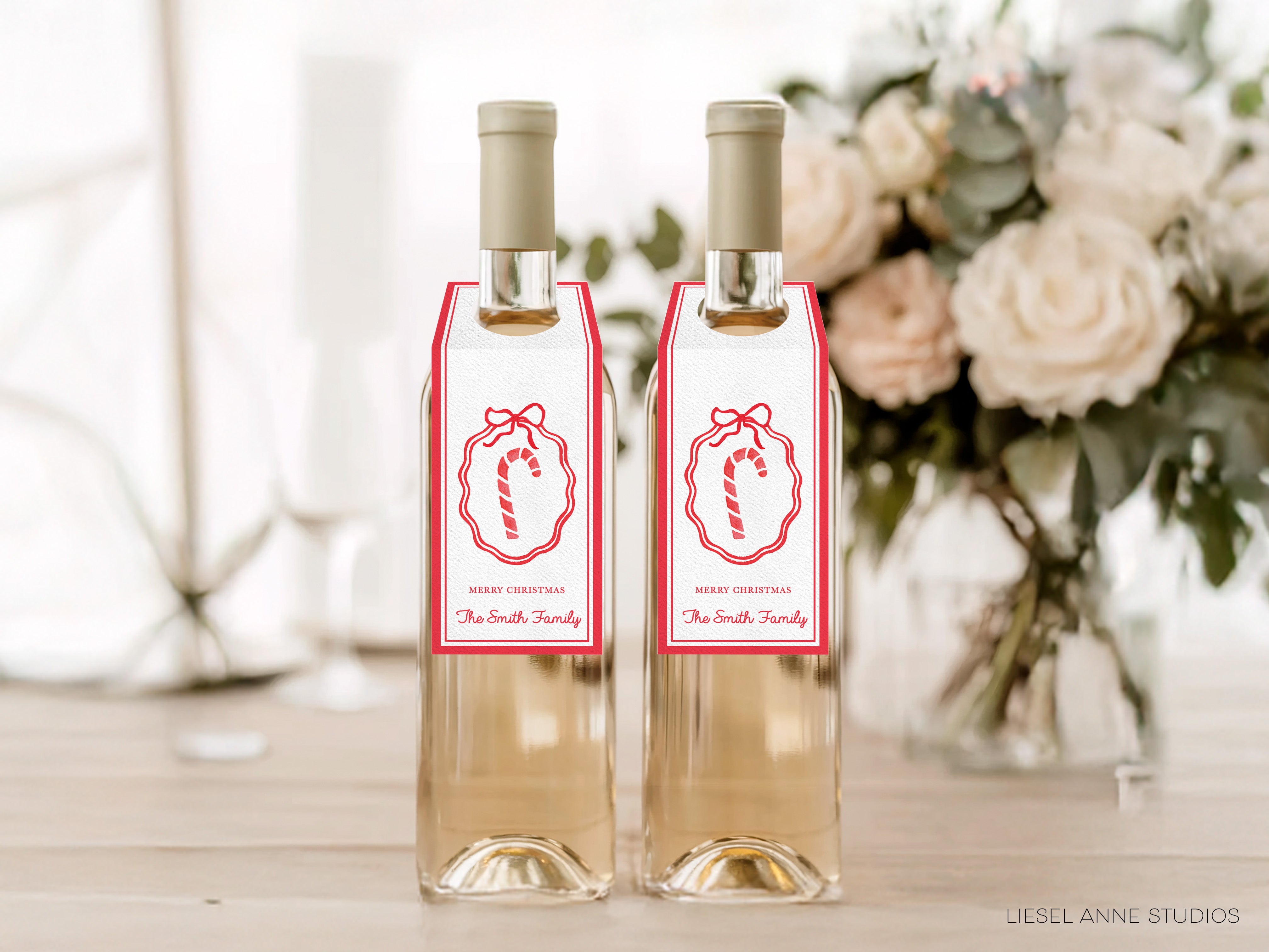 Two bottles of wine with red and white labels, each with a red ribbon and a white ribbon with a red bow, placed on a wooden surface. In the background, there is a vase filled with white flowers.