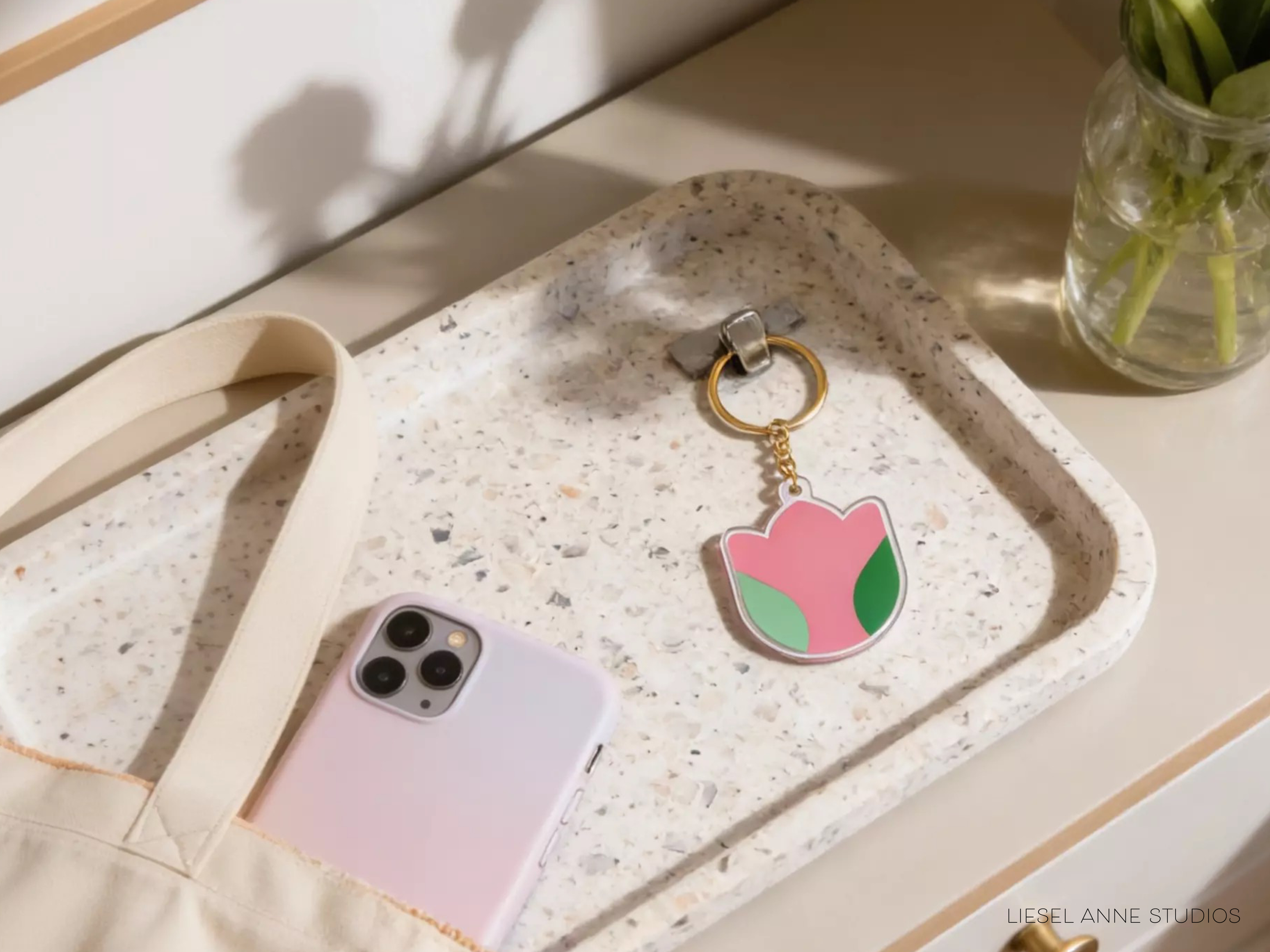 A white tray with a pink and green flower-shaped keychain, a white phone, and a glass vase with green stems on a white surface.