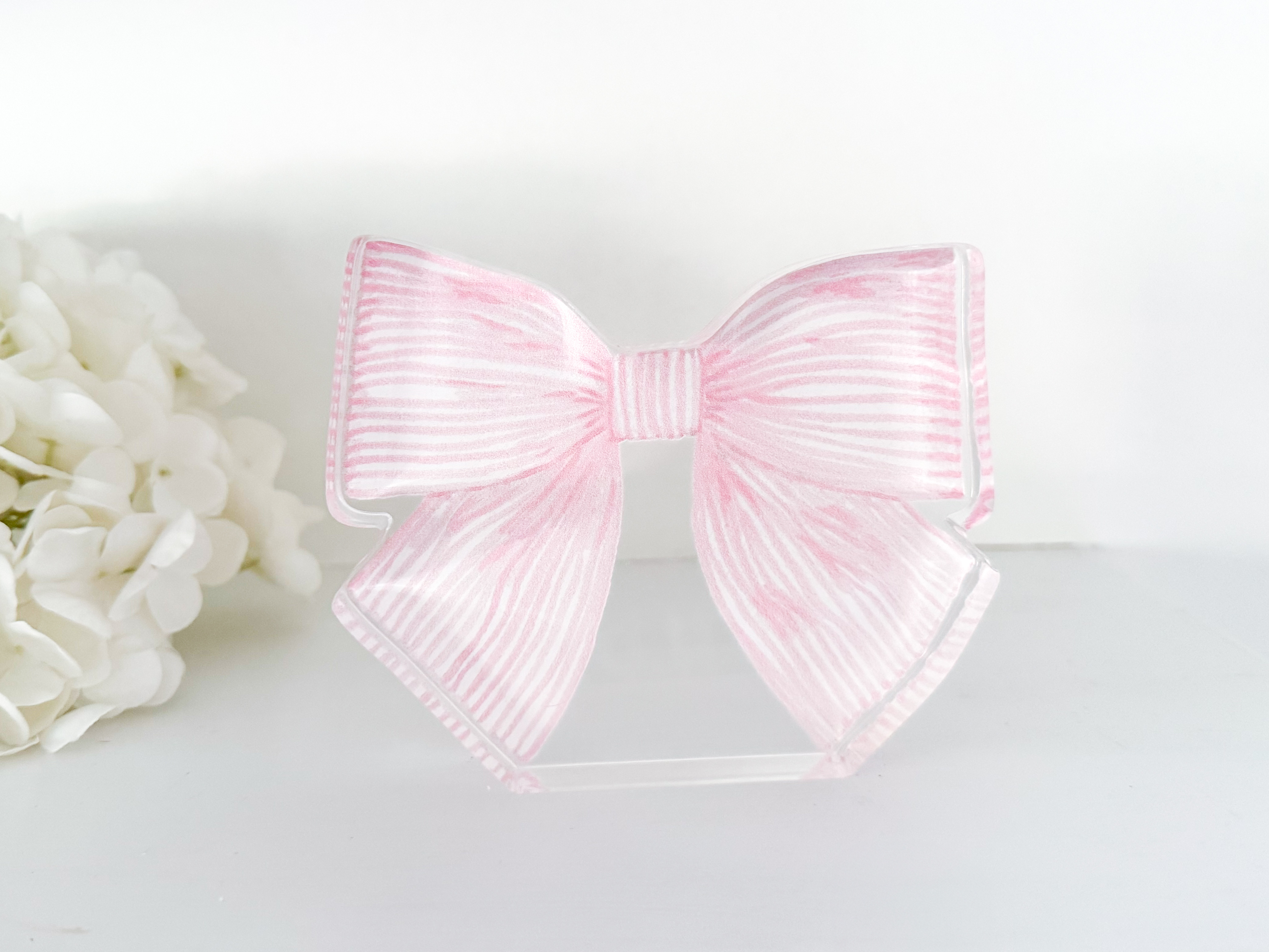A pink and white striped bow is displayed on a white surface, with a bunch of white flowers visible in the background.
