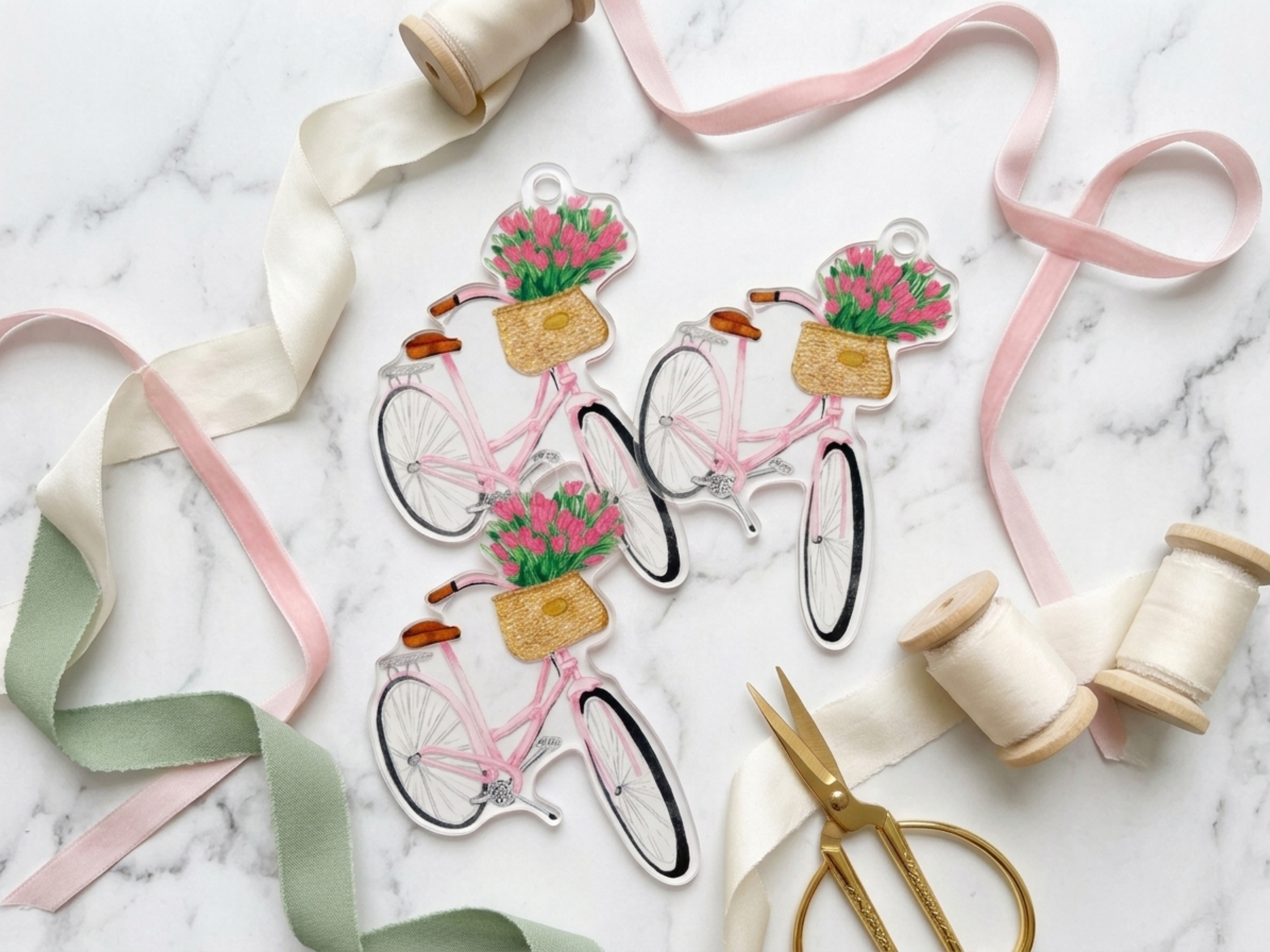 three clear acrylic bicycle charms, each adorned with a basket of pink flowers, arranged on a marble surface alongside various crafting supplies such as ribbon, scissors, and spools of thread.