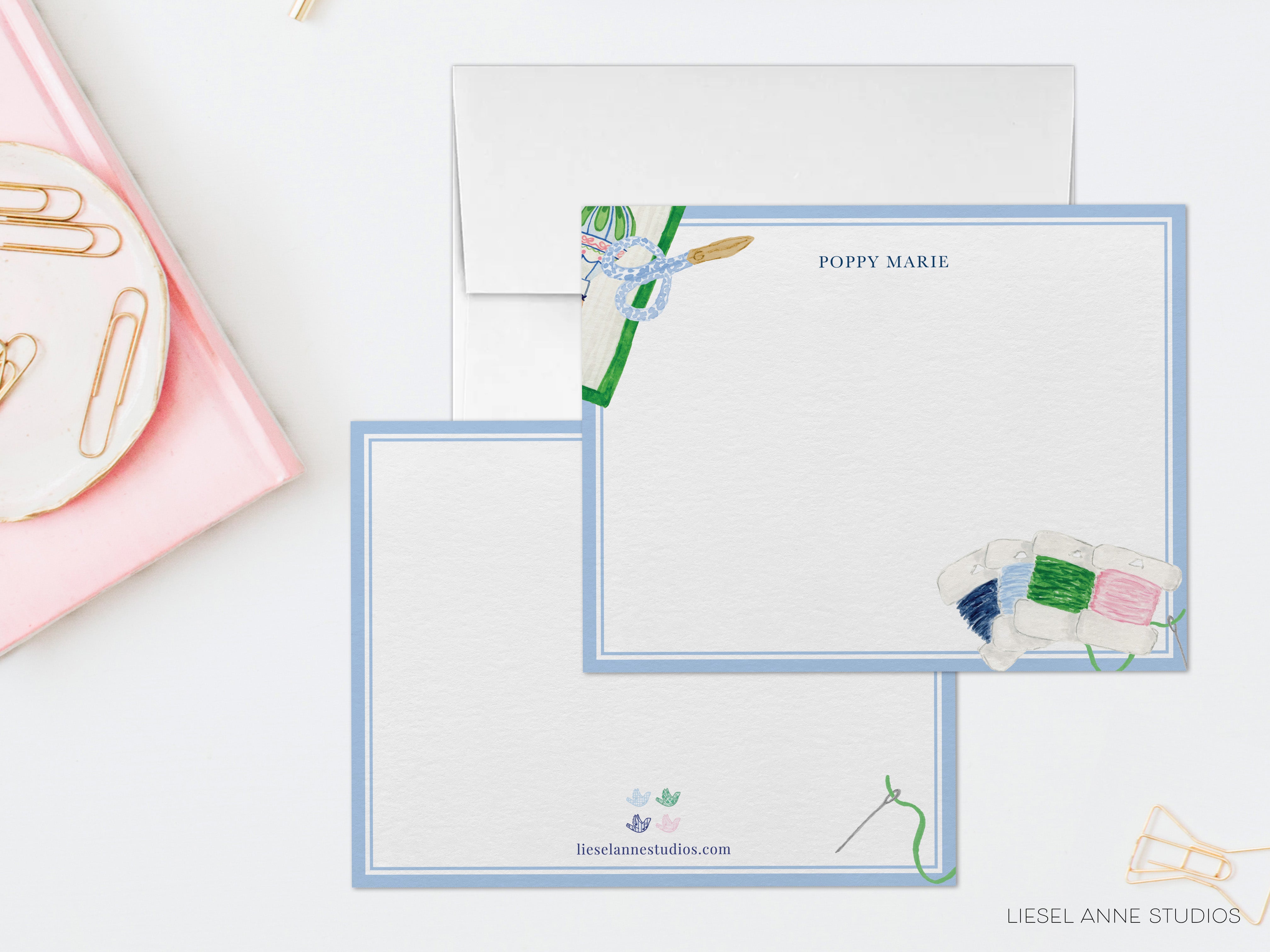 two blank cards with illustrations of sewing supplies, including a needle and thread, and the name "POPPPY MARIE" written on them. The cards are placed on a white surface, with a pink plate and some gold clips visible in the background.