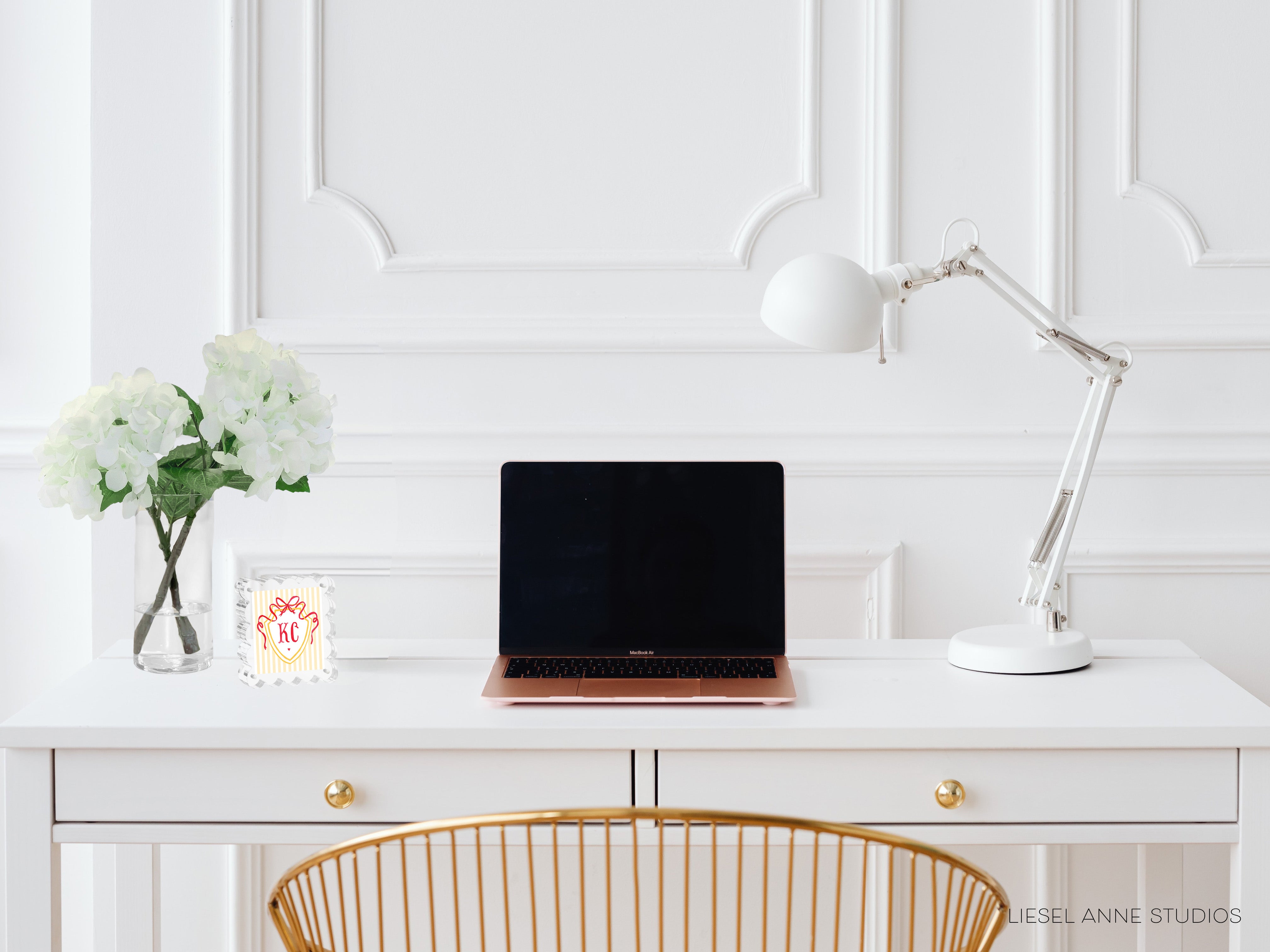 A white desk with a laptop, a lamp, and a vase of white flowers.