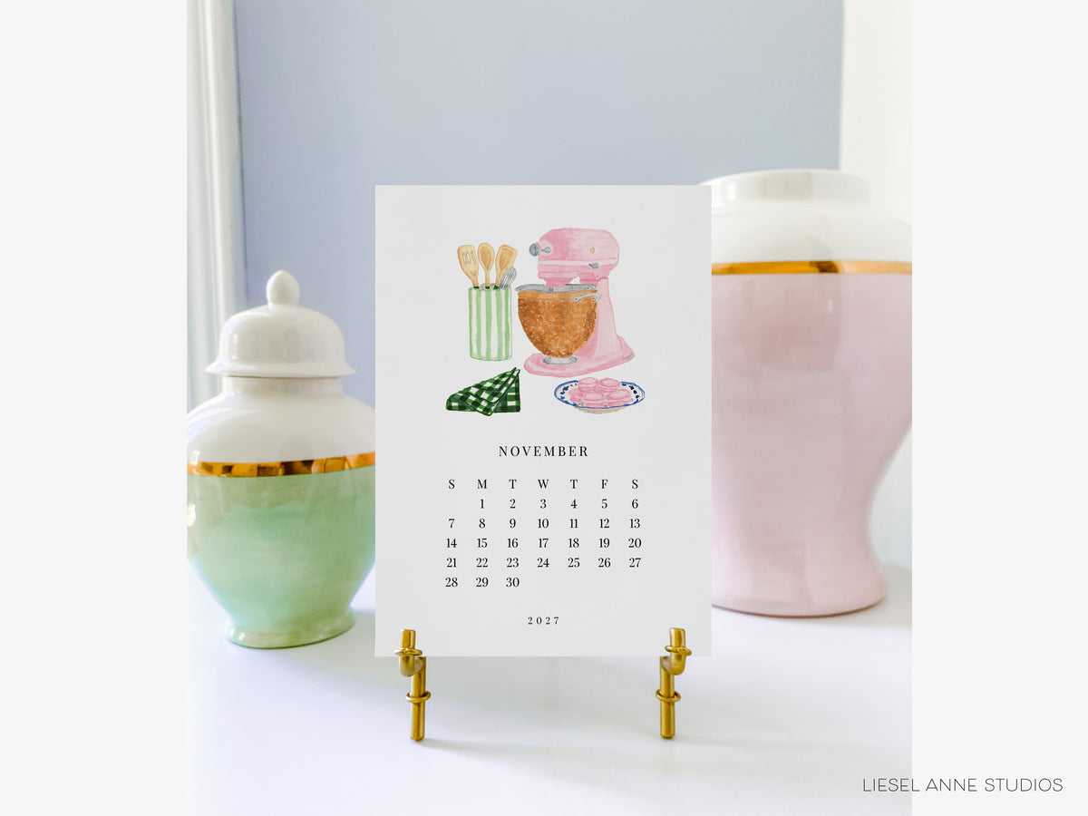 A calendar for the month of November 2017, featuring illustrations of kitchen appliances and utensils, placed on a stand between two decorative vases.