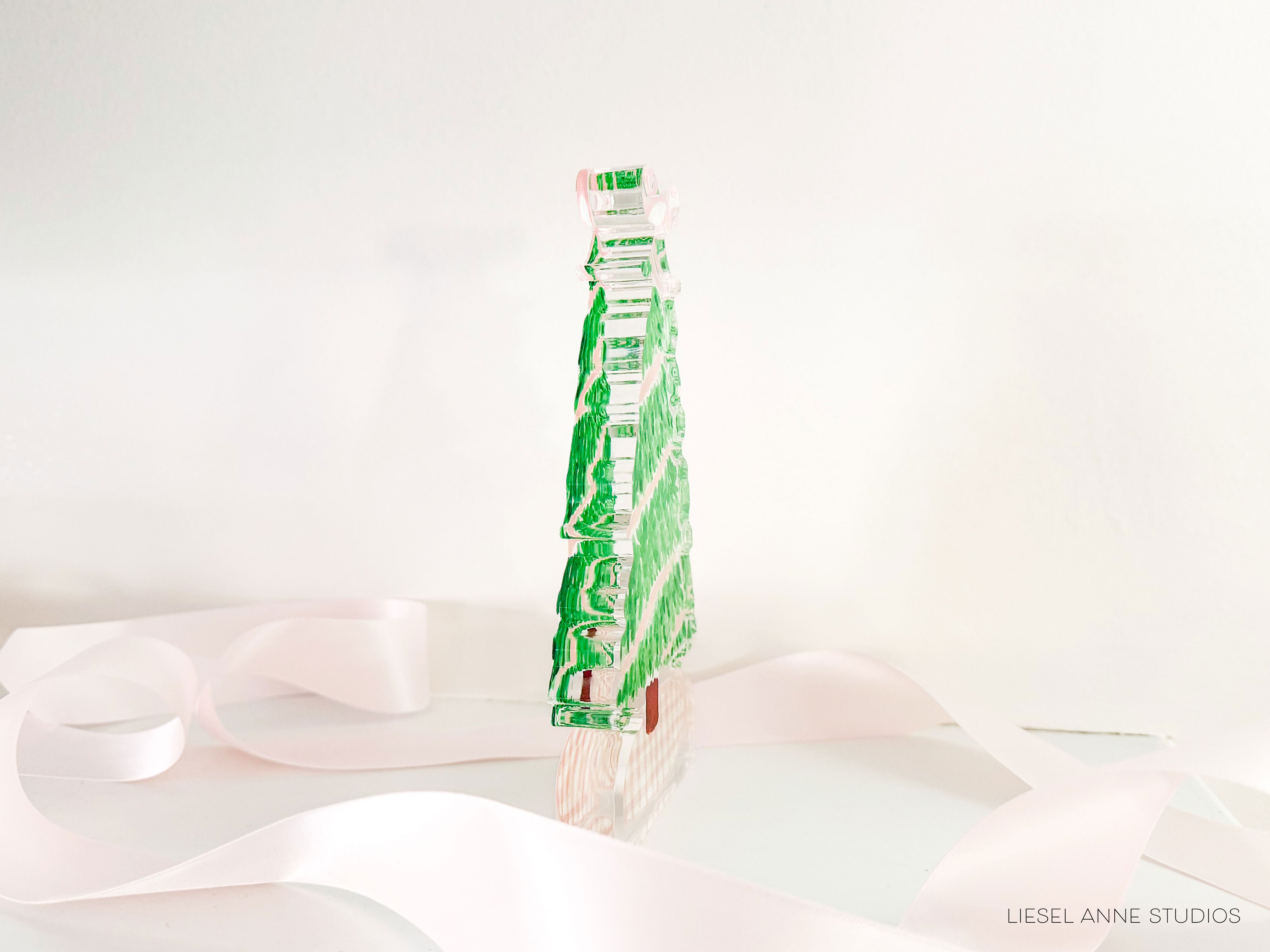 A green glass Christmas tree ornament is displayed on a white background, with a pink ribbon visible in the foreground.