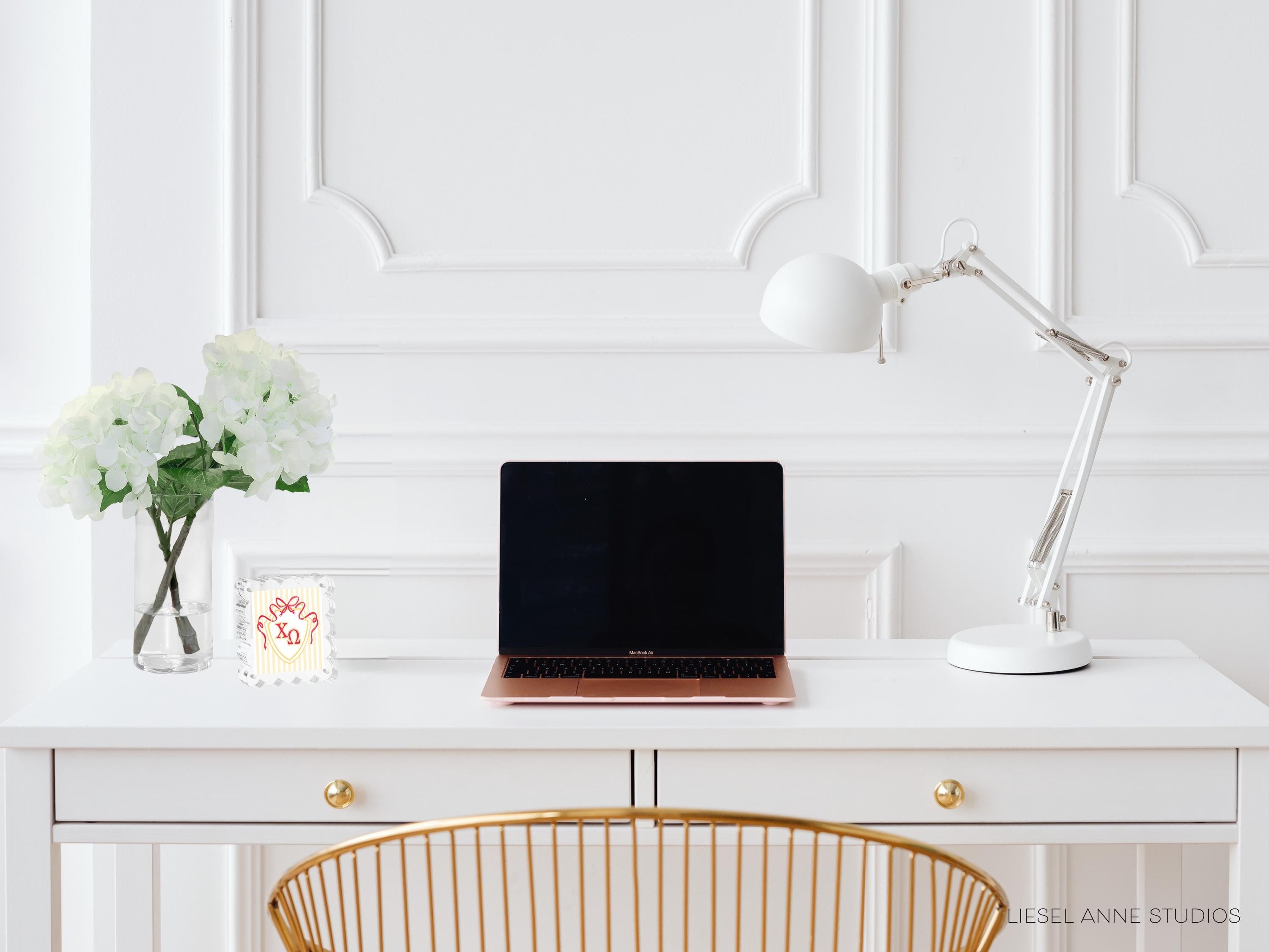 A white desk with a laptop, a lamp, and a vase of white flowers.